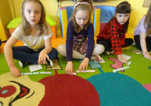 Trzy dziewczynki siedzą na dywanie i układają zdania z rozsypanek wyrazowych.
